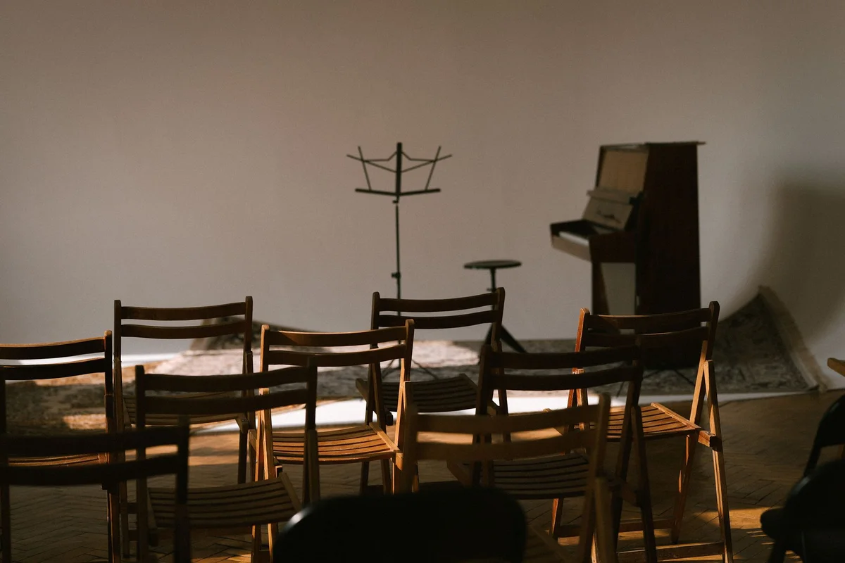 Wooden chairs facing an upright piano and music stand in a small performance room