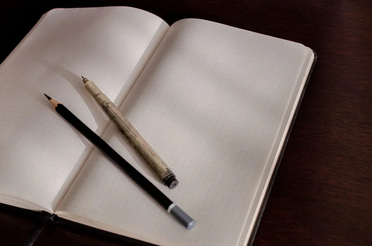 An open dotted notebook with a pencil and pen on a dark wooden desk