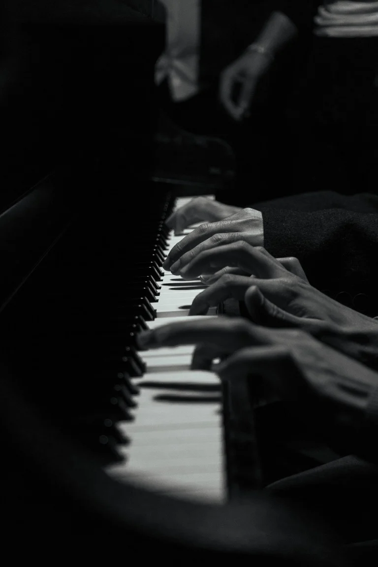 Black-and-white close-up of two players’ hands sharing a piano keyboard