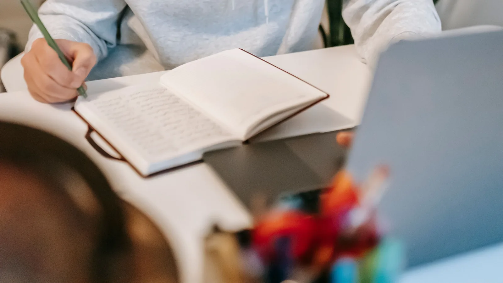 A hand writing in an open notebook beside a laptop