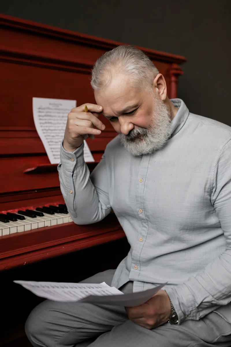 An adult holding a sheet of music beside a red upright piano.